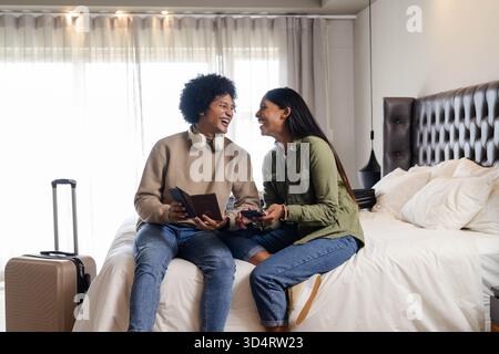Lachende, vielseitige Paare, die auf einem ordentlich gemachten Bett im Hotelzimmer sitzen, mit Koffer und Pässen Stockfoto
