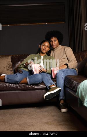 Diverse Paare essen Popcorn aus gestreiften Eimern auf Ledersofa im Wohnzimmer, mit Smartphone Stockfoto