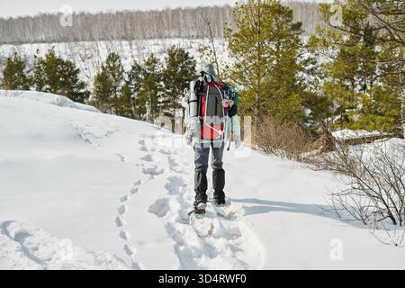Kaukasischer Mann, der durch schneebedeckten Wald mit einem kleinen kaukasischen Kind in einem Rucksackträger wandert, auf einem schneebedeckten Pfad mit sichtbaren Fußspuren, umgeben von einer Winterlandschaft Stockfoto