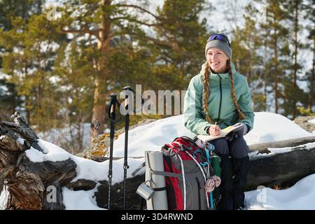 Kaukasische junge Erwachsene Frau, die draußen auf verschneiten Holzfäden sitzt und in Notizbüchern schreibt, lächelt und wegblickt, umgeben von Wanderrucksack und Trekkingstöcken im Winterwald Stockfoto