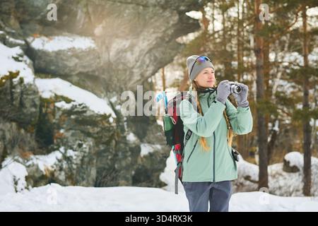 Kaukasische junge Erwachsene Frau, die draußen im verschneiten Wald steht und fotografiert, Rucksack und Winterkleidung trägt, umgeben von Bäumen und Felsen Stockfoto