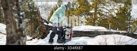 Kaukasischer Mann mittleren Alters, der in verschneiten Wäldern steht und einen großen Wanderrucksack in der Nähe von umgestürzten Bäumen anzieht, Winterkleidung trägt und sich auf das winterliche Trekkingabenteuer vorbereitet Stockfoto