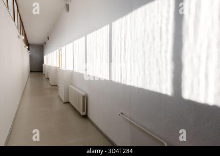 Ein minimalistischer, weißer Flur erstreckt sich bis in die Ferne und ist in natürliches Licht getaucht, das starke vertikale Schatten an der Wand erzeugt. Kühler führen die l Stockfoto