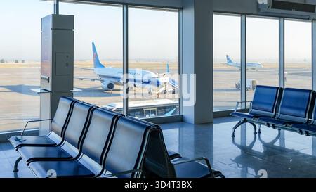 Das Innere des Manas International Airport Terminal mit leerem Sitzbereich und großen Fenstern, die geparkte Flugzeuge auf dem Rollfeld zeigen, schafft eine ruhige Atmosphäre Stockfoto