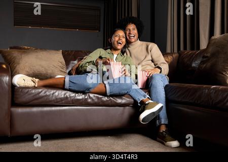 Verschiedene Paare sitzen im Wohnzimmer auf einem Ledersofa und schauen sich Filme an, mit gestreiften Popcorn-Boxen Stockfoto