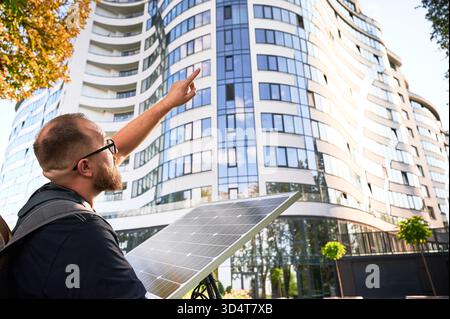Ein Mann, der Photovoltaik-Solarmodul hält, zeigt auf ein gekrümmtes Glasgebäude. Konzept der Integration nachhaltiger erneuerbarer Energiequellen in die moderne Architektur. Stockfoto