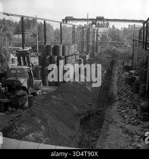 Ein schwarzweißes Archivfoto aus den 1970er Jahren zeigt ein groß angelegtes Bau- oder Erweiterungsprojekt im Öllager Sloviansk, der ukrainischen SSR. Die Szene wird von einem tiefen, langen Graben dominiert, der in die Erde gegraben wurde und wahrscheinlich für die Installation neuer Rohre mit großem Durchmesser geeignet ist. Neben der Ausgrabung stapeln sich Hunderte von Metallfässern in massiven, hohen Säulen auf dem Lagerplatz. Links steht ein Schwerlaststapler, und über dem gesamten Standort läuft ein Portalkransystem. Dieses Bild zeigt eindrucksvoll die schweren Bauarbeiten und Erdarbeiten, die beim Bau der industriellen Infrastruktur von erforderlich sind Stockfoto