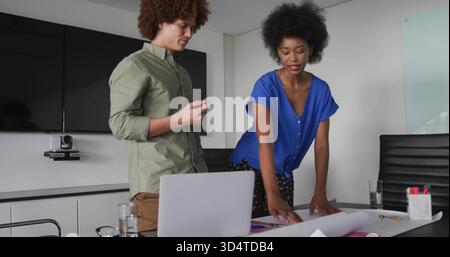 Zeigende Frau in blauer Bluse und Mann in olivem Hemd, die Karten im Büro überprüft, mit Laptop Stockfoto