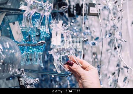 Hand hält ein durchsichtiges Rentier-Ornament in einem Ladengang voller glitzernder Weihnachtsdekoration. Nachhaltiges Weihnachtsgeschäft, umweltfreundlich Stockfoto