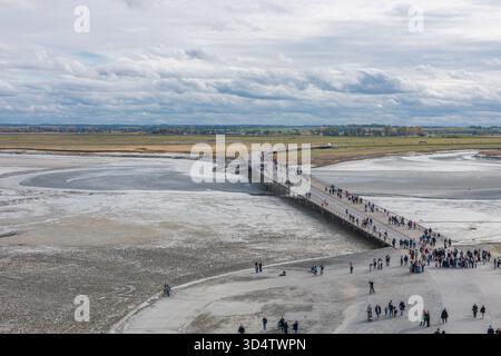 Panoramablick vom Mont Saint-Michel aus mit felsigen Wänden, geparkten Fahrzeugen und Besuchern, die auf den exponierten Wattflächen unter einem bewölkten Himmel in niedriger Tiefe spazieren Stockfoto