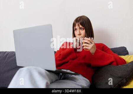Die junge Frau entspannt sich zu Hause, konzentriert sich auf ihren Laptop, während sie einen warmen Drink schlürft. Stockfoto