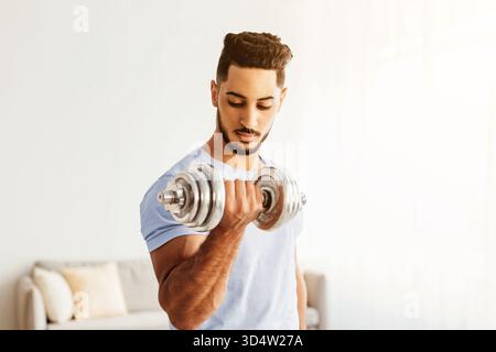 Junger Mann, der Hantel in einem hellen Raum hebt und für Fitness und Kraft trainiert Stockfoto