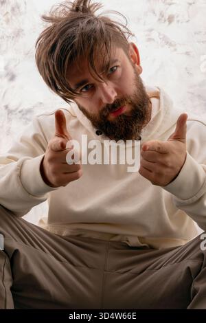 Junger bärtiger Mann in lässigem Hoodie, der selbstbewusst auf die Kamera zeigt und Positivität und freundliche Einstellung vermittelt. Stockfoto
