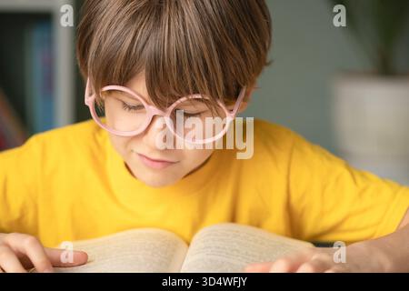 Ein lächelnder Schüler in einer Brille liegt die Hand, während er ein offenes Buch am Schulpult liest. Kindliches Lernen, kluger Geist, Studienkonzept, Frühentwicklung und Vorbereitung auf Bildung und künftiges Wachstum. Stockfoto