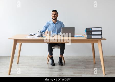Lächelnder arabischer Mann, der die Arbeit im Home Office mit Laptop und Kaffee in der Hand genießt Stockfoto