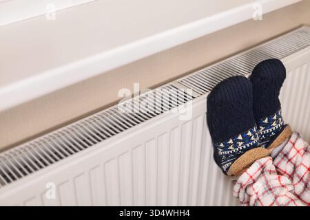 Bequeme Füße in warmen Wollsocken und kariertem Pyjama auf einem weißen Heizkörper für winterlichen Komfort. Hochwertige Fotos Stockfoto
