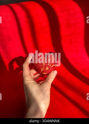 Eine Hand hält sanft ein leuchtendes rotes, knackiges, herzförmiges Choux-Gebäck vor einem auffälligen roten Hintergrund mit dramatischen Schatten. Dieses süße Leckerli Sy Stockfoto