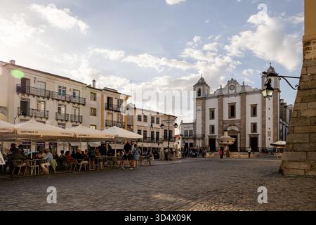 Malerischer Blick auf Praca do Giraldo in Evora, Portugal während der goldenen Stunde, mit Einheimischen und Touristen, die Cafés im Freien unter großen Sonnenschirmen genießen Stockfoto