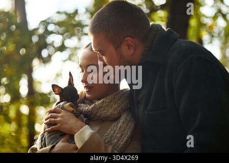 Kaukasische junge Erwachsene Frau und Mann, die im Freien einen kleinen Hund halten, beide sanft das Haustier berühren und das Tier mit Zuneigung betrachten, während sie im Herbstwald spazieren gehen Stockfoto