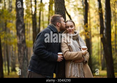 Kaukasischer junger erwachsener Mann, der die Stirn einer Freundin küsst, die eine Kaffeetasse hält, zusammen im Herbstwald steht. Beide tragen warme Kleidung und wirken liebevoll Stockfoto