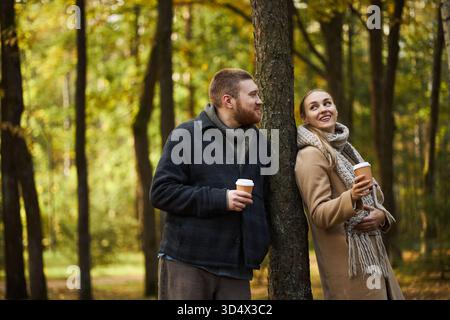 Kaukasischer junger Mann und kaukasische junge Frau, die im Herbstwald stehen und Kaffeetassen zum Mitnehmen halten, sich gegen einen Baum lehnen, lächeln und einander anschauen, in warmer Kleidung Stockfoto
