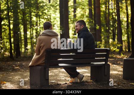 Junger kaukasischer Mann lächelt und hält eine Kaffeetasse, während er auf der Parkbank sitzt und mit einer jungen kaukasischen Frau spricht, Hintergrund im Herbst mit Sonnenlicht, das durch Bäume filtert Stockfoto