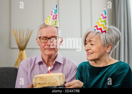 Wünschen Sie sich etwas. Familie Seniorenpaar in Partymütze feiert gemeinsam Geburtstag zu Hause. Ein alter Mann, der Kerzen auf der Geburtstagskuche brennt. Alte Frau wünscht Ehemann alles Gute zum Geburtstag || Model veröffentlicht Stockfoto
