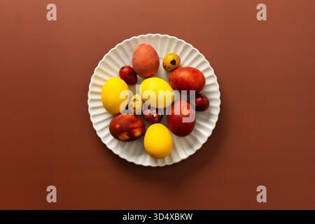 Eine Sammlung von bunten Ostereiern in verschiedenen Größen und Mustern wird auf einer weißen, gewellten Platte auf einer warmen braunen Oberfläche ausgestellt. Stockfoto