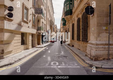 Valletta, Malta, Altstadt, enge Straße, historisches Gebäude, mediterrane Architektur, Sandsteinfassaden, traditioneller Holzbalkone, maltesische Hauptstadt, Stadtgasse, urbanes Leben, Fußgänger, Geschäftshaus, Straßenszene, Alltag in Valletta, Architektur Valletta, historische Altstadt, typisches Stadtbild, kulturelles Erbe, UNESCO-Welterbe, Stadt Malta, mediterrane Atmosphäre, alte Bausubstanz, urbanes Malta, Stadtzentrum, Reiseimpression, Architekturdetails, Steinmauern, Stadtansicht, Straßenperspektive, Altstadtgasse, touristische Region, maltesische Kultur, Hauptstadt Valletta, Stadtarchite Stockfoto