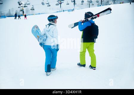 Zwei Freunde stehen im Schnee und machen sich auf Ski- und Snowboardfahrten in einem Bergresort vorbereitet. Der Himmel ist bewölkt und die Pisten sind voller Wintersportarten Stockfoto