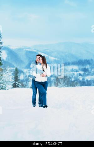 Ein Paar steht im Schnee und umschließt sich warm, während es an einem Wintertag von atemberaubenden Bergen unter einem klaren blauen Himmel umgeben ist. Stockfoto