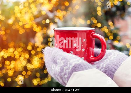 Weibliche Hände in kuscheligen Strickhandschuhen, die eine rote Tasse mit einem heißen Getränk, Kaffee oder Glühwein vor einem Weihnachtsbaum halten Stockfoto
