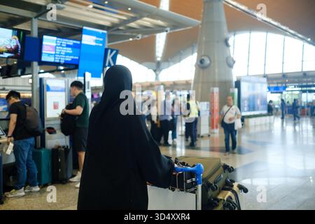 Eine Frau in einem schwarzen Hijab schiebt einen Gepäckwagen an einem Flughafen. Andere Reisende werden beim Check-in und beim Blick auf Fluginformationstafeln im bac gesehen Stockfoto