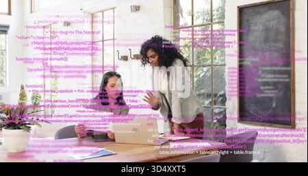 Kooperierende Frauen in Blazer und dunklem Top, die sich im Startup-Büro über einen Laptop lehnen, mit Code-Overlay Stockfoto