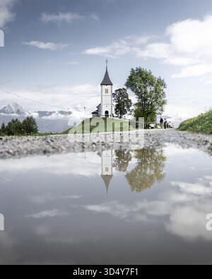Blick auf eine Kirche auf einem Hügel, die sich in einer Pfütze auf einem Kiesweg spiegelt, Jamnik, Slowenien. Stockfoto