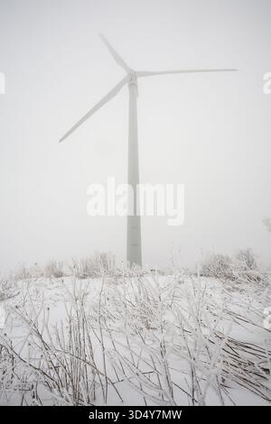 Der Blick auf eine einsame Windturbine erhebt sich durch eine dicke Nebeldecke, deren Blätter kaum sichtbar sind, vor der gedämpften Winterlandschaft in Syracuse, New York Stockfoto