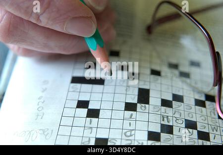 Eine Frau füllt vorsichtig ein Kreuzworträtsel mit einem grünen Bleistift aus. Das Puzzle erscheint in einer Zeitung und ihre Brille liegt auf der Seite neben ihr. Stockfoto
