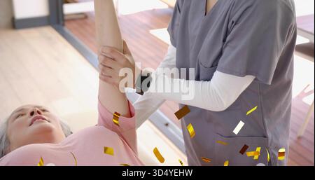Betreuerin in grauen Schutten hält Seniorin mit rosa Oberseite auf dem Tisch im Therapiesaal, Smartwatch Stockfoto