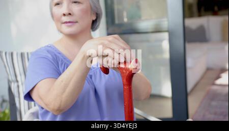 Sitzende ältere asiatische Frau mit Lavendeloberteil, die roten Stock auf der Veranda mit gestreiftem Kissen hält Stockfoto