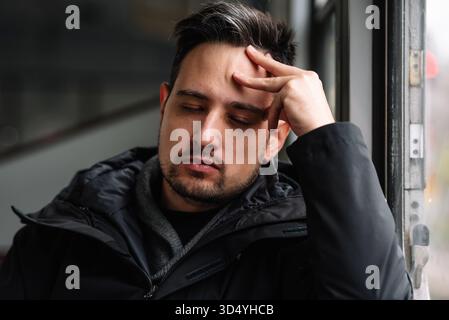 Müder junger Mann mit geschlossenen Augen, der Kopf auf der Hand am Fenster liegt Stockfoto