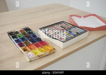 Auf einem Holztisch werden zwei Kunstgegenstände ausgestellt. Auf der linken Seite befindet sich neben einer Schachtel mit Pastelltönen eine lebhafte Aquarellpalette mit vielen Farben. Perfekt für Stockfoto