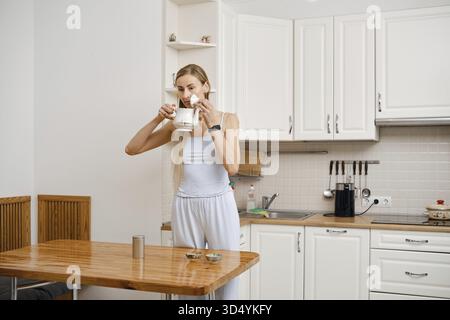 In einer modernen Küche atmet eine junge Frau in leichter Kleidung das Aroma von frisch gebrühtem Tee aus einer Teekanne ein. Weiches Morgenlicht erfüllt den Raum, während sie Stockfoto