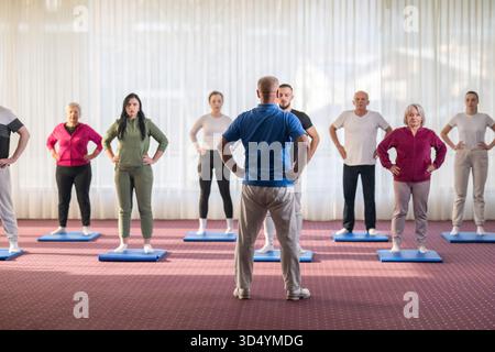 Trainer führt eine vielfältige Gruppe von Menschen während eines Fitnesstrainings mit Dehnungsübungen auf Matten in einem hellen Wellnesscenter Stockfoto