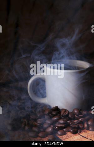 Dampfender heißer Kaffee in einem weißen Becher, umgeben von verstreuten gerösteten Bohnen auf einer dunklen rustikalen Holzoberfläche, warmes und aromatisches Ambiente Stockfoto