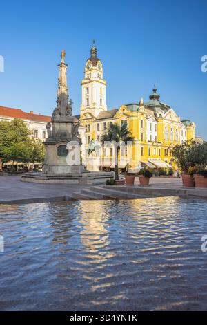 Pecs, Ungarn - 13. Juli 2024: Szechenyi-Platz. Der kunstvoll verzierte europäische platz mit Uhrenturm, religiösem Denkmal und reflektierendem Pool unter einem klaren Himmel Stockfoto