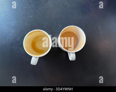 Eine Aufnahme von zwei weißen Keramikbechern mit Restkaffee- oder Teeflecken auf einer glatten, dunklen Oberfläche. Stockfoto