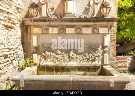 Granada, Spanien - 5. September 2024: Alhambra-Komplex, reich verzierter Steinbrunnen mit skulpturalen Wassertieren, vor einer alten Ziegelmauer. Wasserdurchfluss Stockfoto
