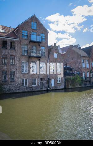 Historische Backsteingebäude säumen einen ruhigen Kanal in Gent, Belgien, mit Reflexionen im grünlichen Wasser, modernen Glasbalkonen und einem hellblauen Himmel Stockfoto