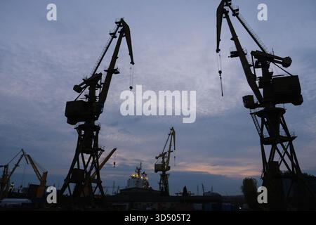 Silhouetten großer Industriekräne stehen vor dem Abendhimmel in einem Hafengebiet. Eine dramatische Szene, die Bauwesen, Industrie, Logistik usw. symbolisiert Stockfoto