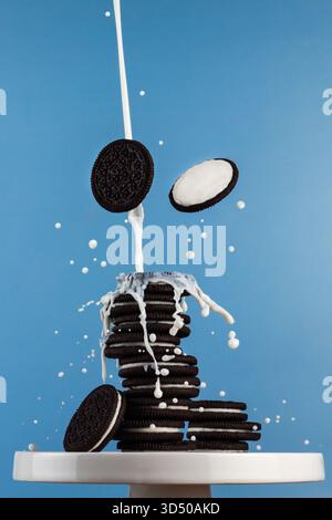 Kekse fallen aus einem hohen Stapel, während Milch in einem verspielten Dessertmoment vor einem hellblauen Hintergrund in der Küche nach oben spritzt. Stockfoto